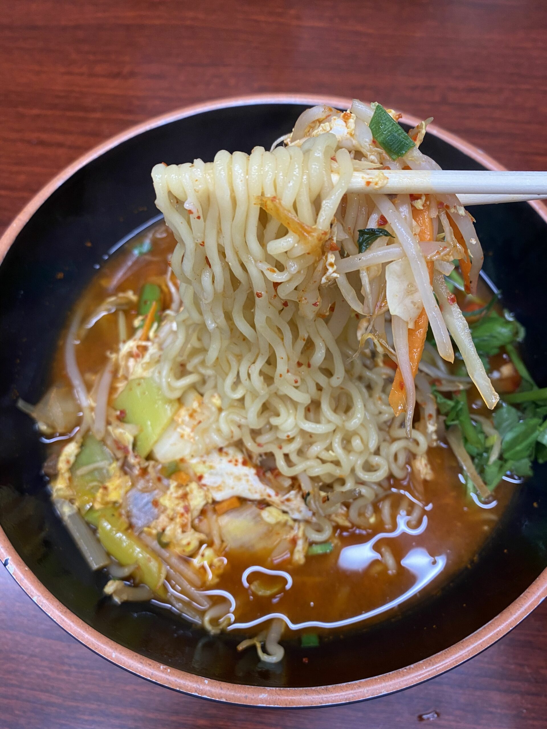 A bowl of spicy broth, Korean veggies and ramen noodles being held up with chopsticks